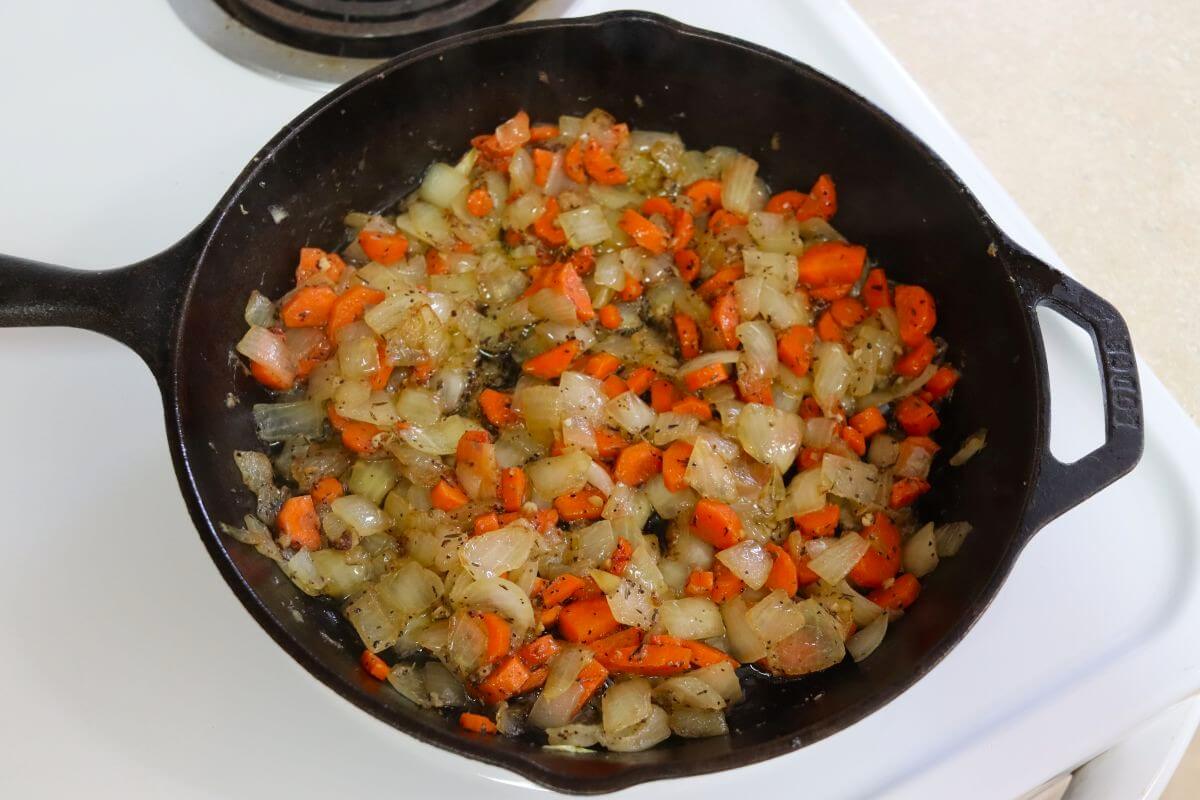 Diced carrots and onions cooking in butter with garlic and herbs in a cast iron skillet on the stovetop.