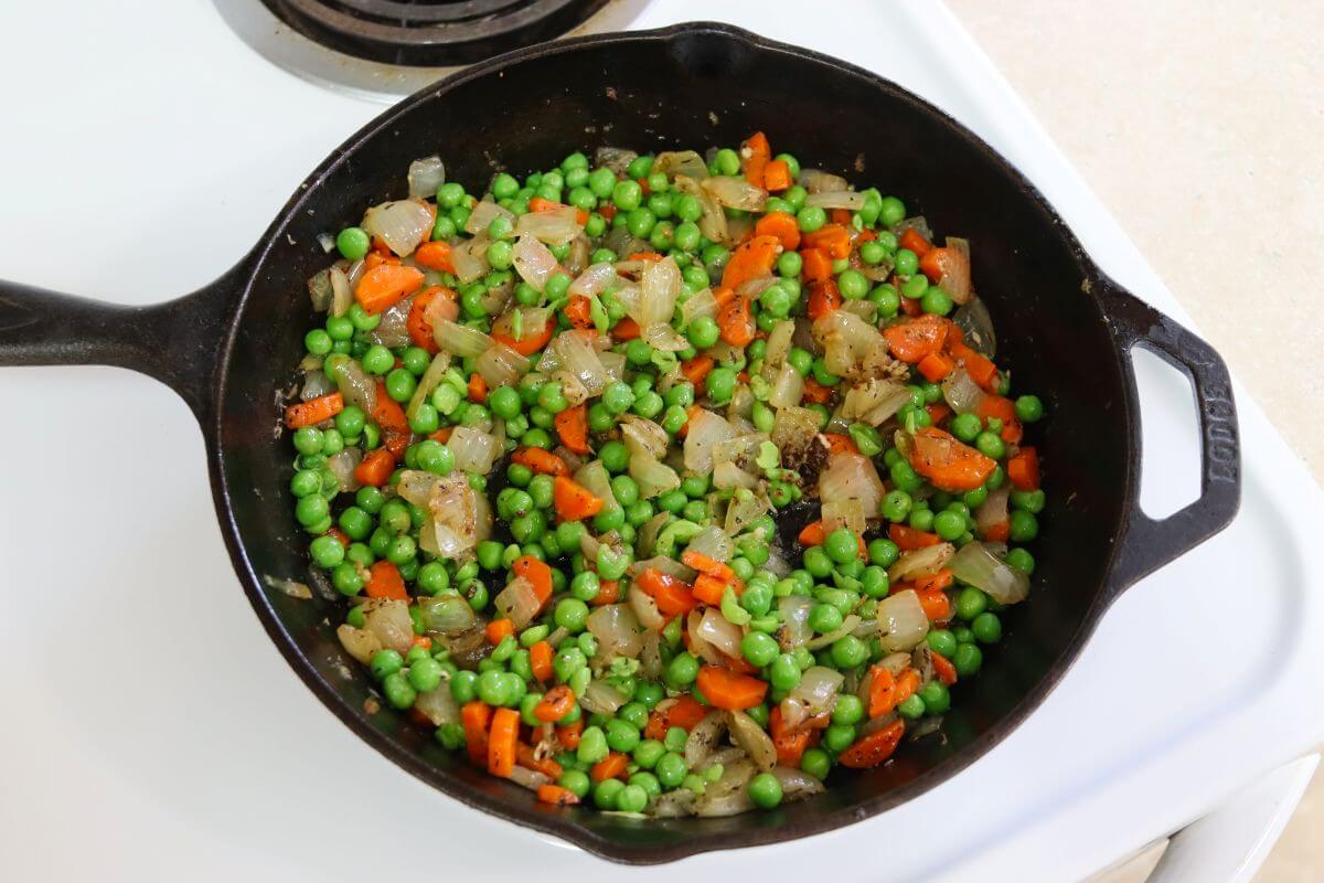 Peas added to cooked carrots and onions in a cast iron skillet on the stovetop.