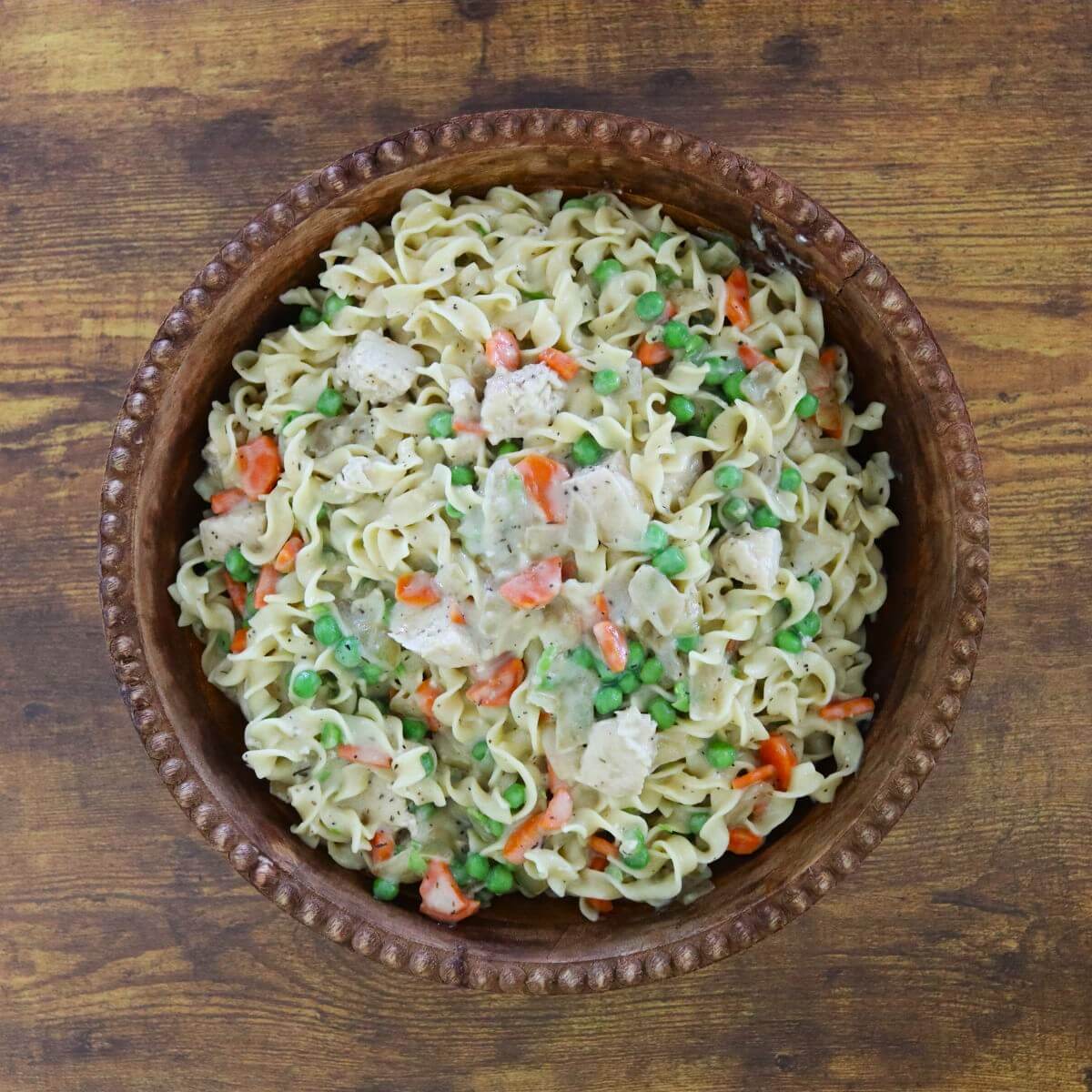 Full bowl of creamy chicken and noodles showing egg noodles mixed with chicken, carrots, and peas in a thick sauce.
