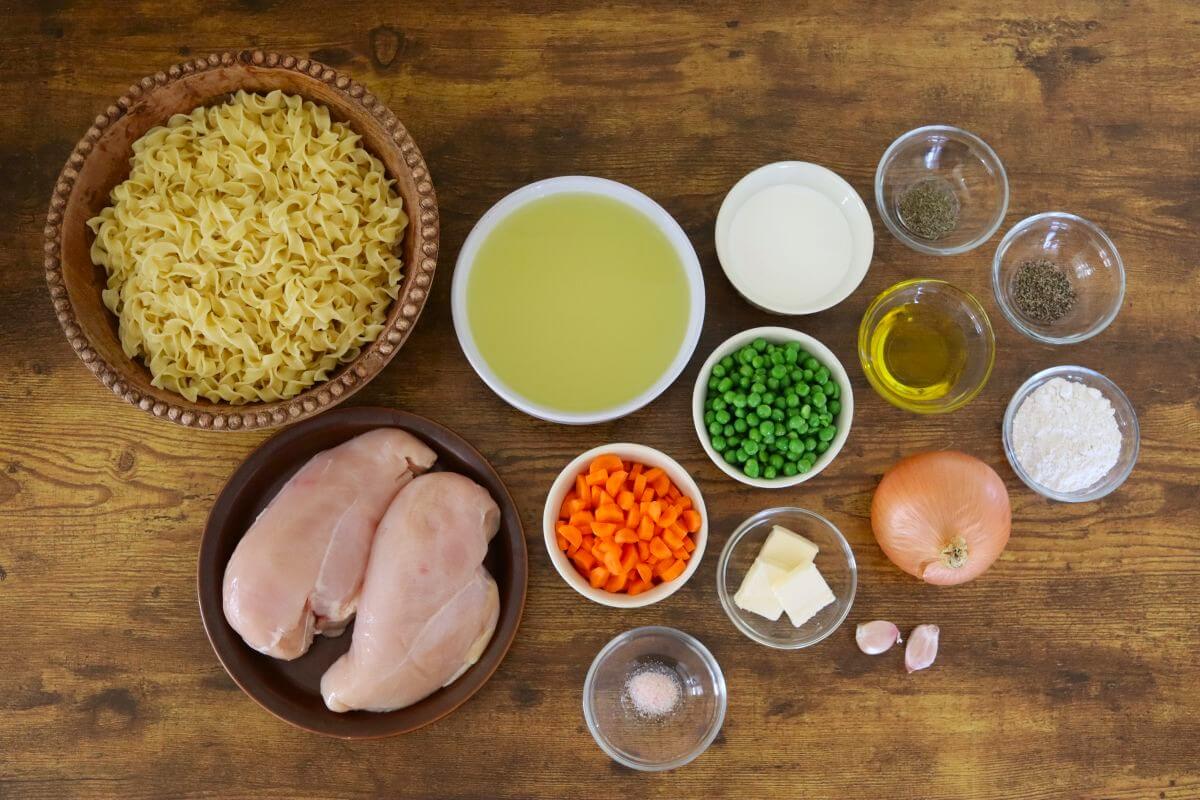Ingredients for chicken and noodles arranged on a wooden table, including chicken breast, egg noodles, chicken broth, cream, peas, carrots, onion, garlic, butter, olive oil, flour, salt, pepper, and thyme.