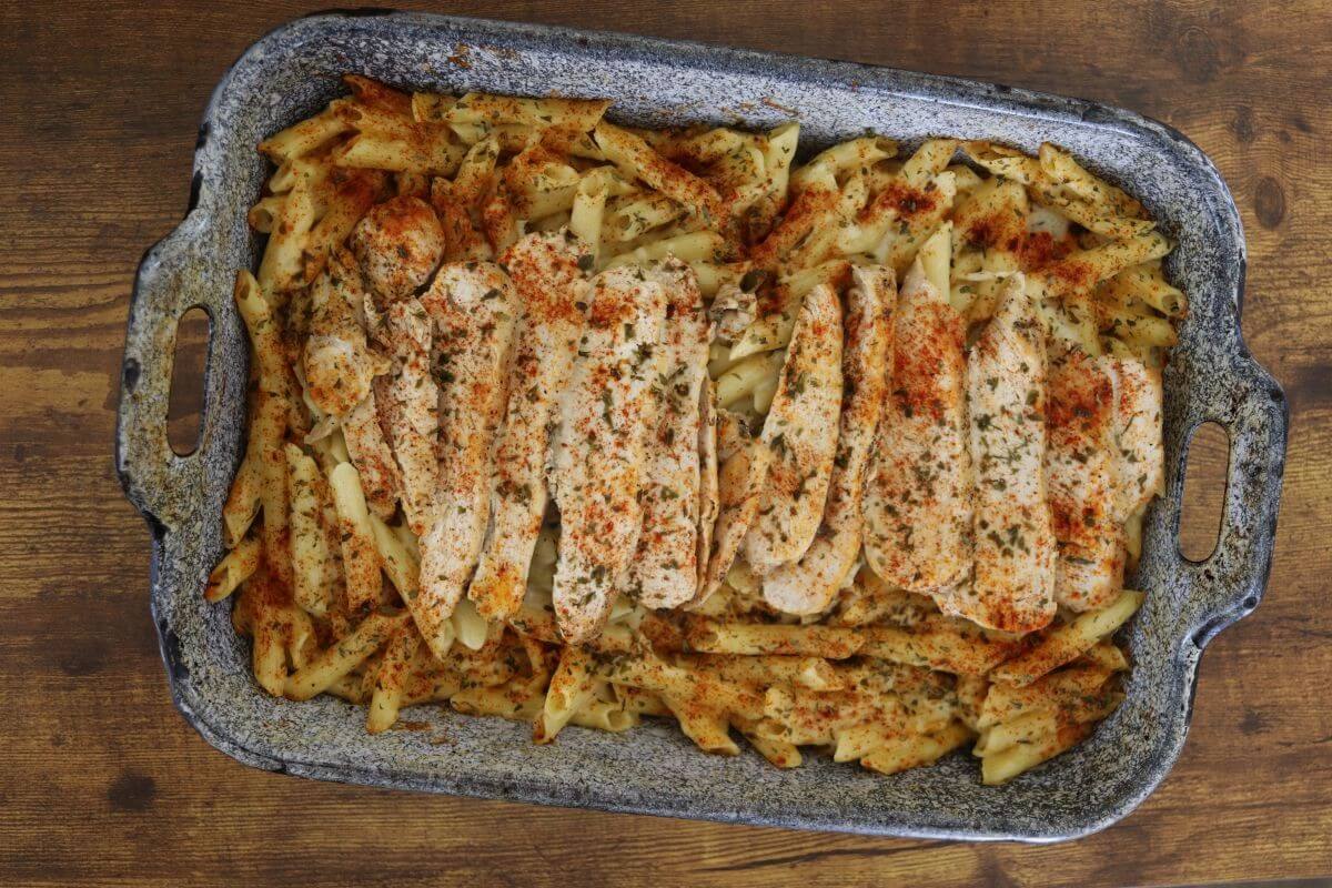 Baked penne pasta in a creamy white sauce with sliced chicken breast, parsley, and paprika sprinkled on top in a baking pan.
