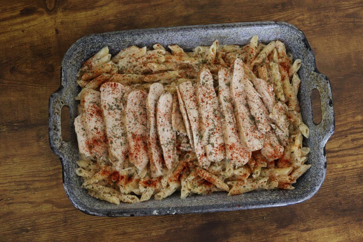 Penne pasta in a creamy white sauce with sliced chicken breast, parsley, and paprika sprinkled on top in a baking pan.