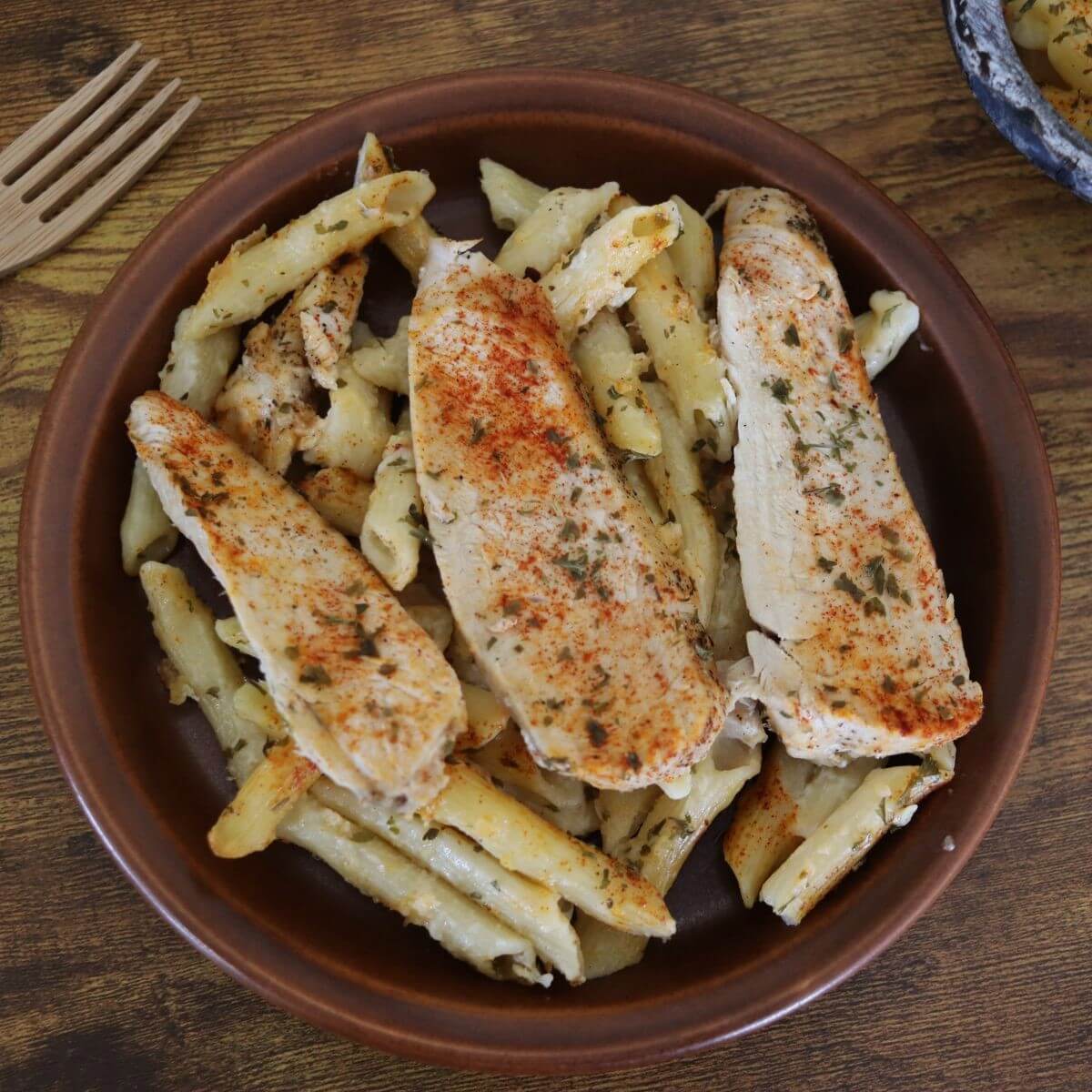 Penne pasta in white cream sauce topped with slices of chicken breast, parsley, and paprika on a plate.