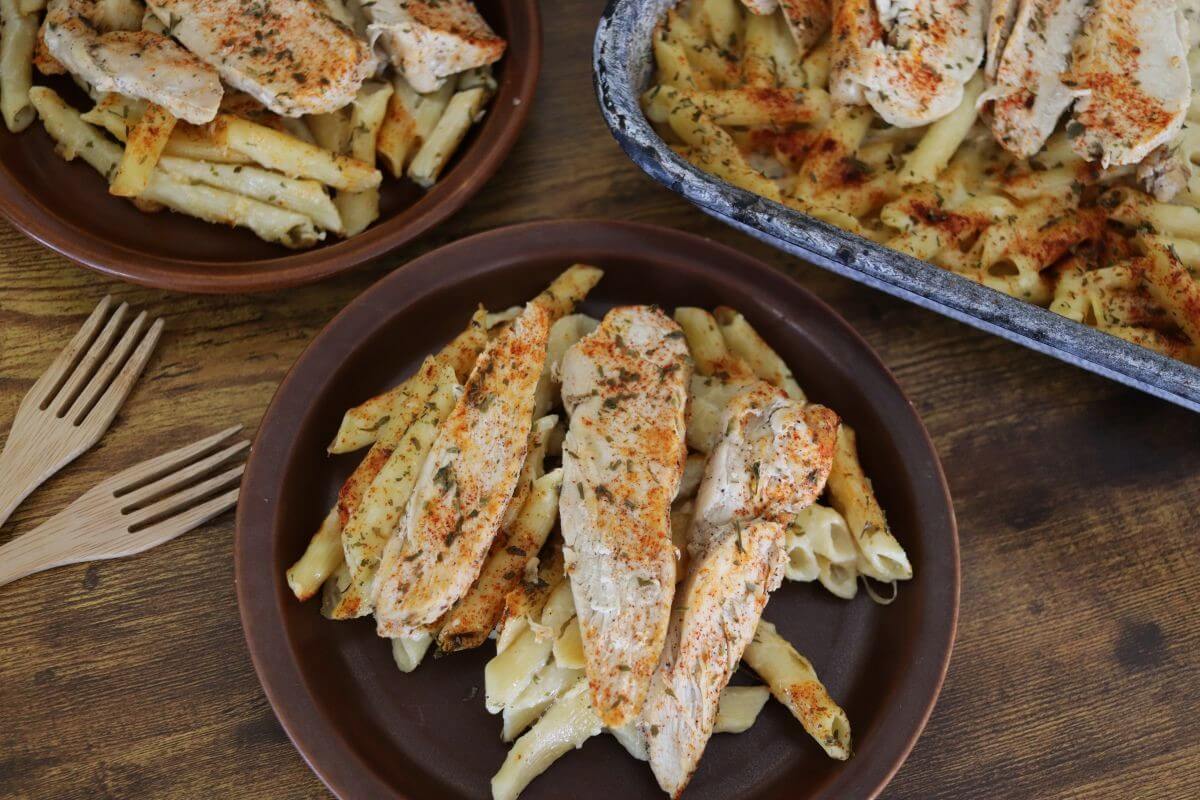 A plate of baked penne pasta topped with seasoned grilled chicken strips, served on a brown plate with another serving and a baking dish in the background, alongside two wooden forks on a wooden table.