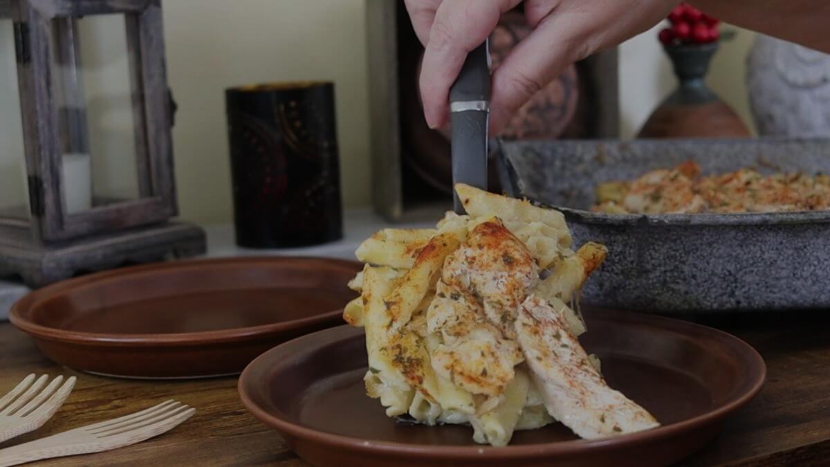 A hand serves a portion of baked pasta with pieces of chicken onto a brown plate. The dish is cheesy and golden, set on a wooden table with a tray of more pasta, wooden forks, and decorative items in the background.
