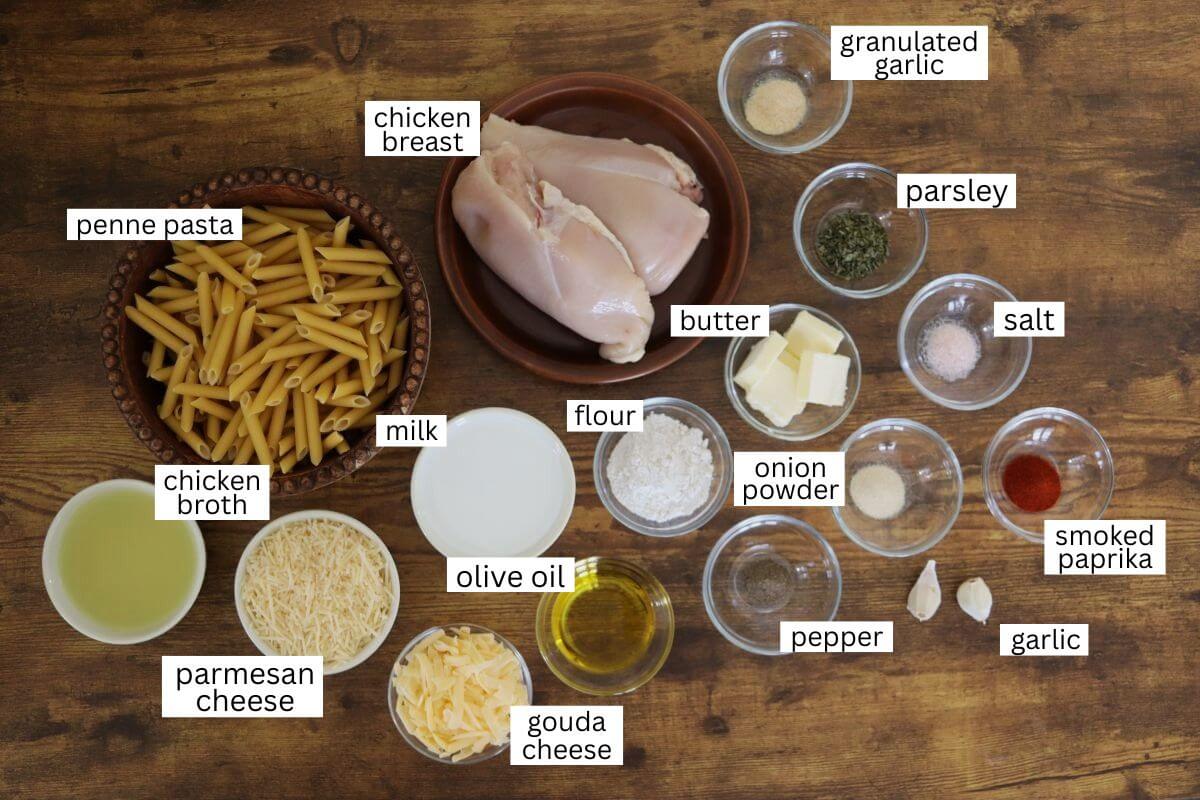 A variety of ingredients for a chicken pasta dish displayed on a wooden table, including raw chicken breast, penne pasta, cheeses, broth, milk, spices, olive oil, butter, flour, and minced garlic in labeled bowls.