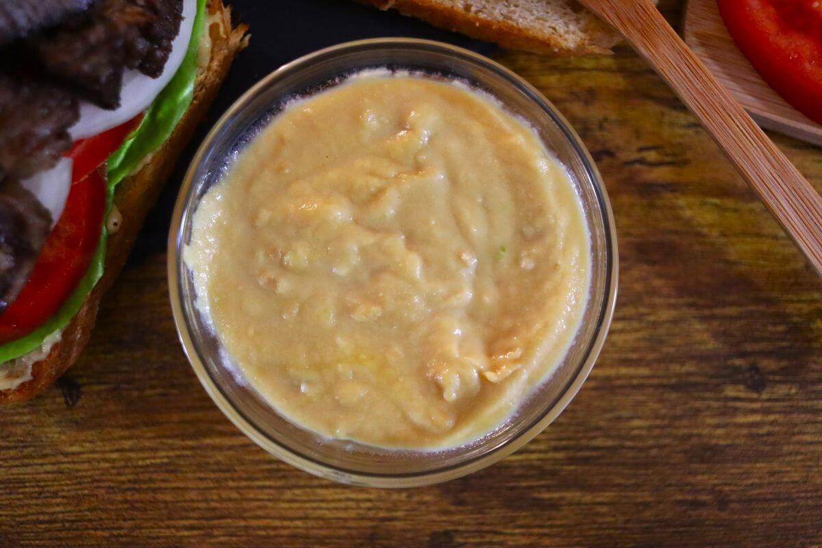 A small glass bowl of creamy, light tan sauce sits on a wooden surface next to sandwich ingredients, including lettuce, tomato, and bread.