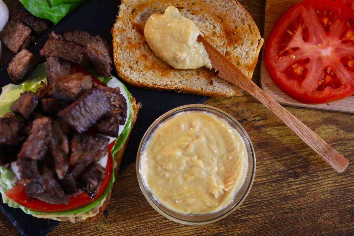 An open-faced sandwich with sliced tomato, lettuce, and grilled steak pieces sits next to a slice of bread being spread with a creamy sauce. A bowl of the same sauce and more tomato slices are nearby on a wooden surface.