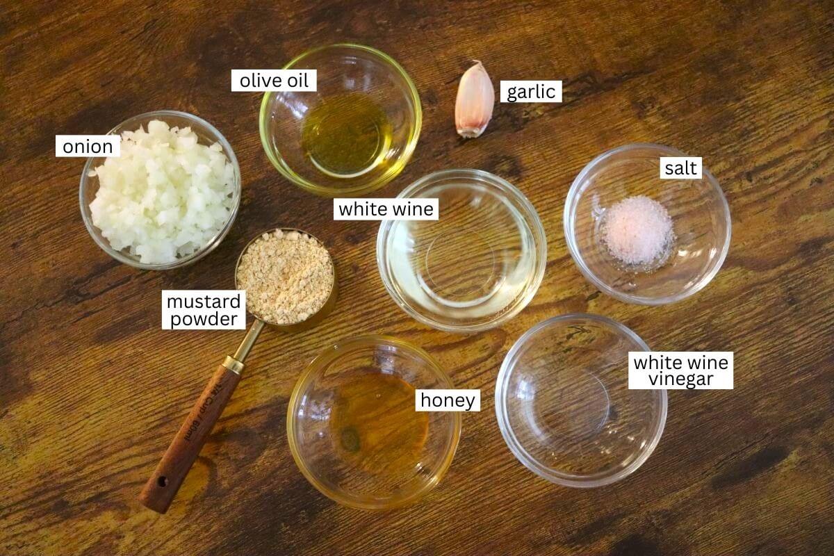 A wooden table holds bowls with labeled ingredients: chopped onion, olive oil, a garlic clove, white wine, salt, mustard powder, honey, and white wine vinegar, arranged in a casual layout.