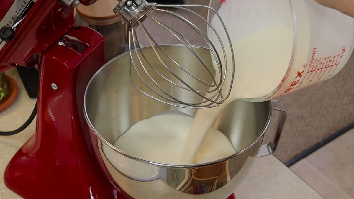 Heavy cream being poured into a stainless steel mixing bowl with a large whisk visible above it.
