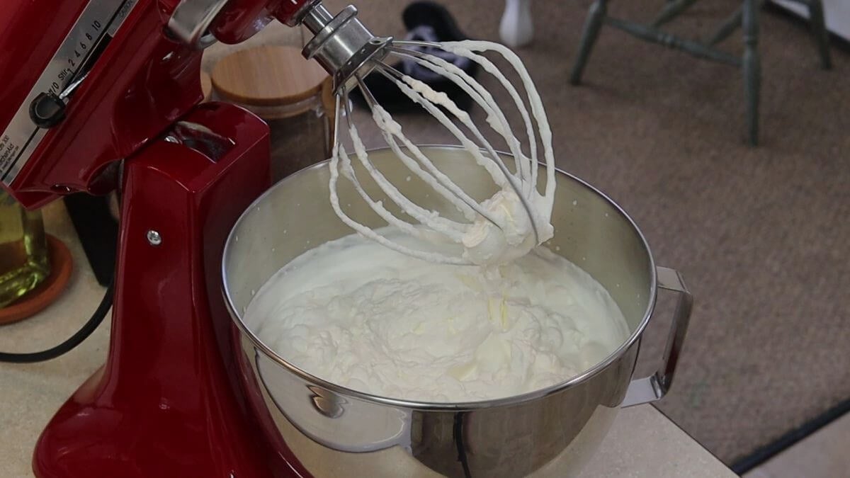 The stainless steel bowl of a stand mixer full of whipped cream with the whisk attachment, covered in whipped cream, above it.