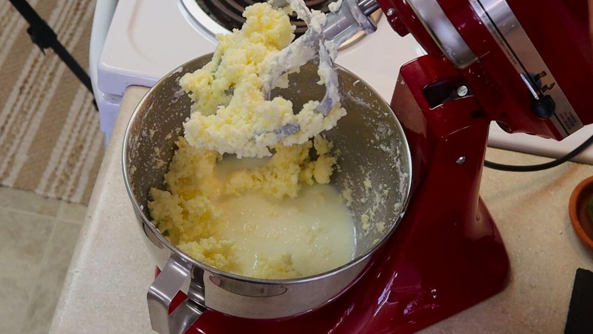 Butter and buttermilk in the bowl of a stand mixer with the paddle attachment, covered in butter, above it.