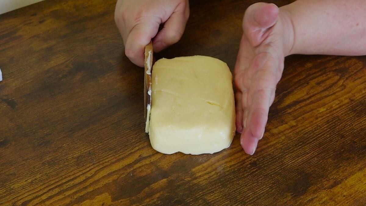 A hand pushing a wooden paddle against the side of a rectangle of butter, with another hand pushing on the other side.