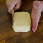 Two hands shaping a chunk of homemade butter into a rectangle, one with a wooden paddle in it.