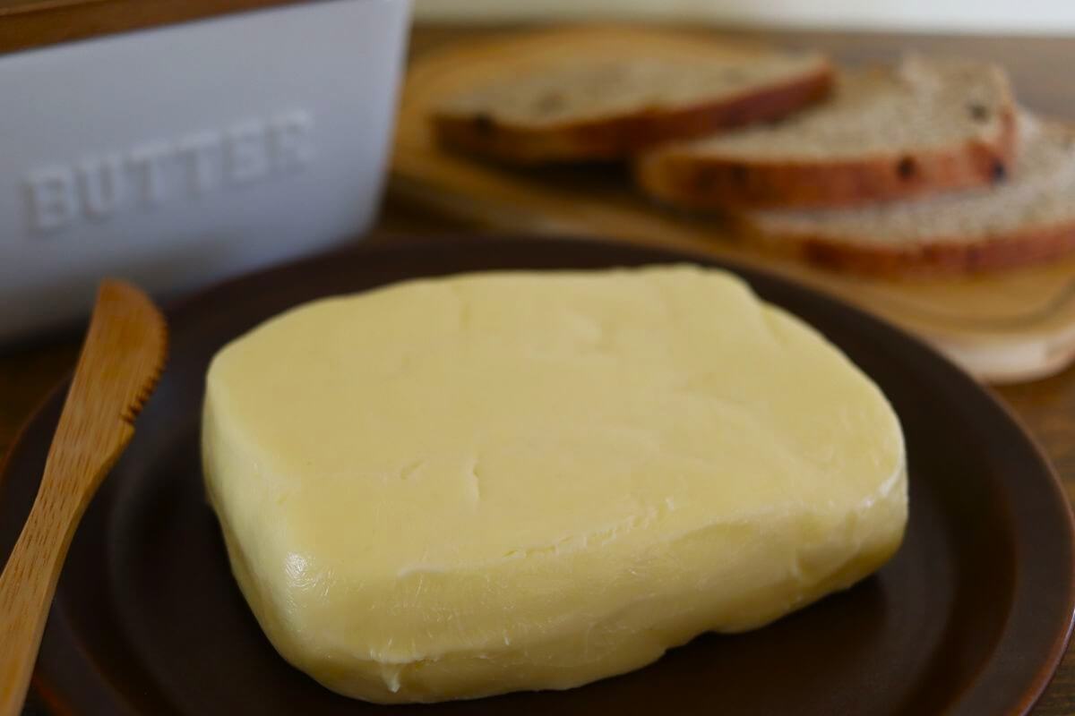 A block of butter sits on a brown plate next to a wooden knife, with slices of bread and a butter container in the background.