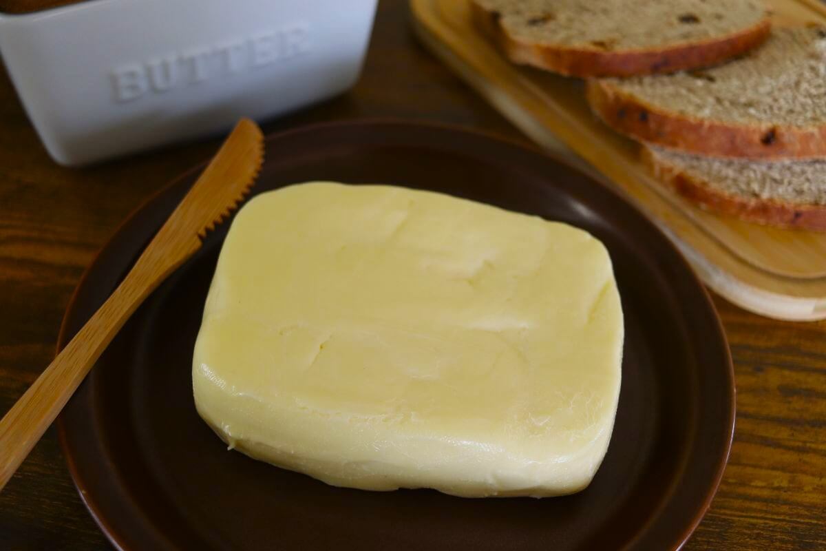 A rectangular block of butter on a brown plate with a wooden butter knife beside it. Slices of bread and a butter dish labeled “BUTTER” are in the background on a wooden table.