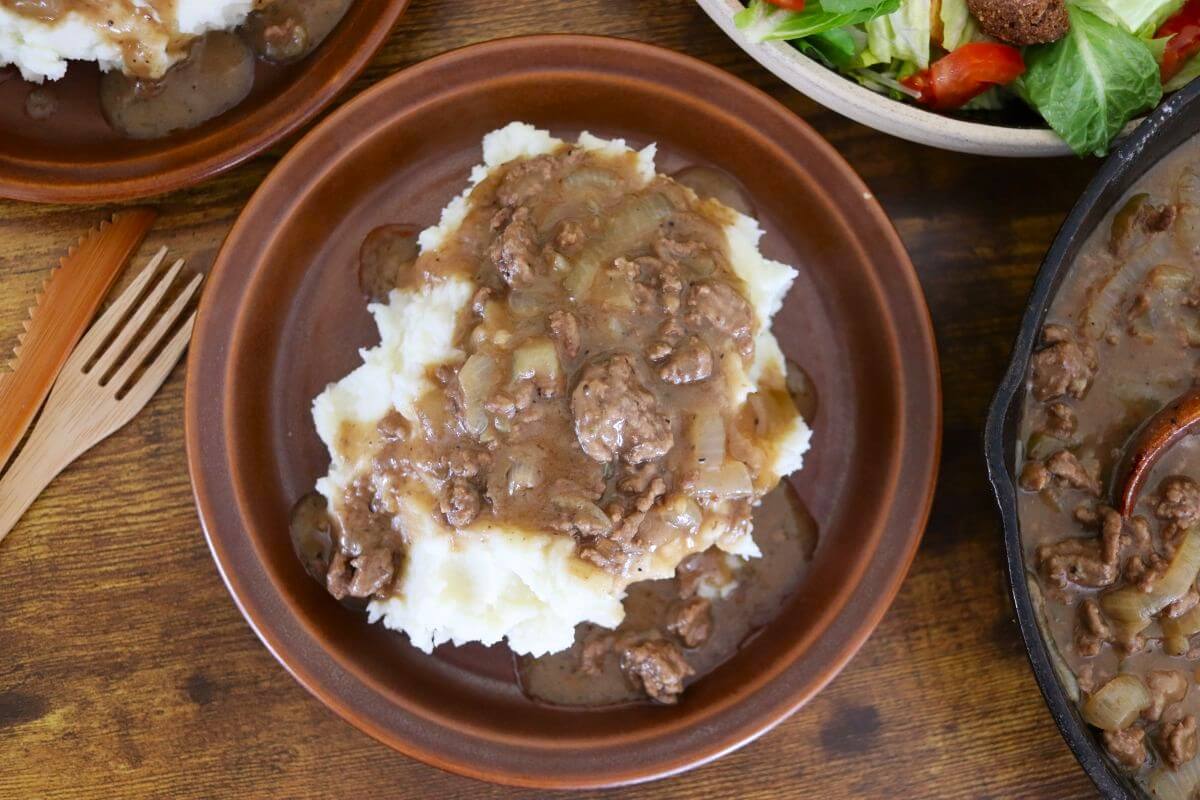 Old-fashioned ground beef and gravy served over mashed potatoes on a brown plate, with a cast iron skillet of gravy beside it on a wooden table.