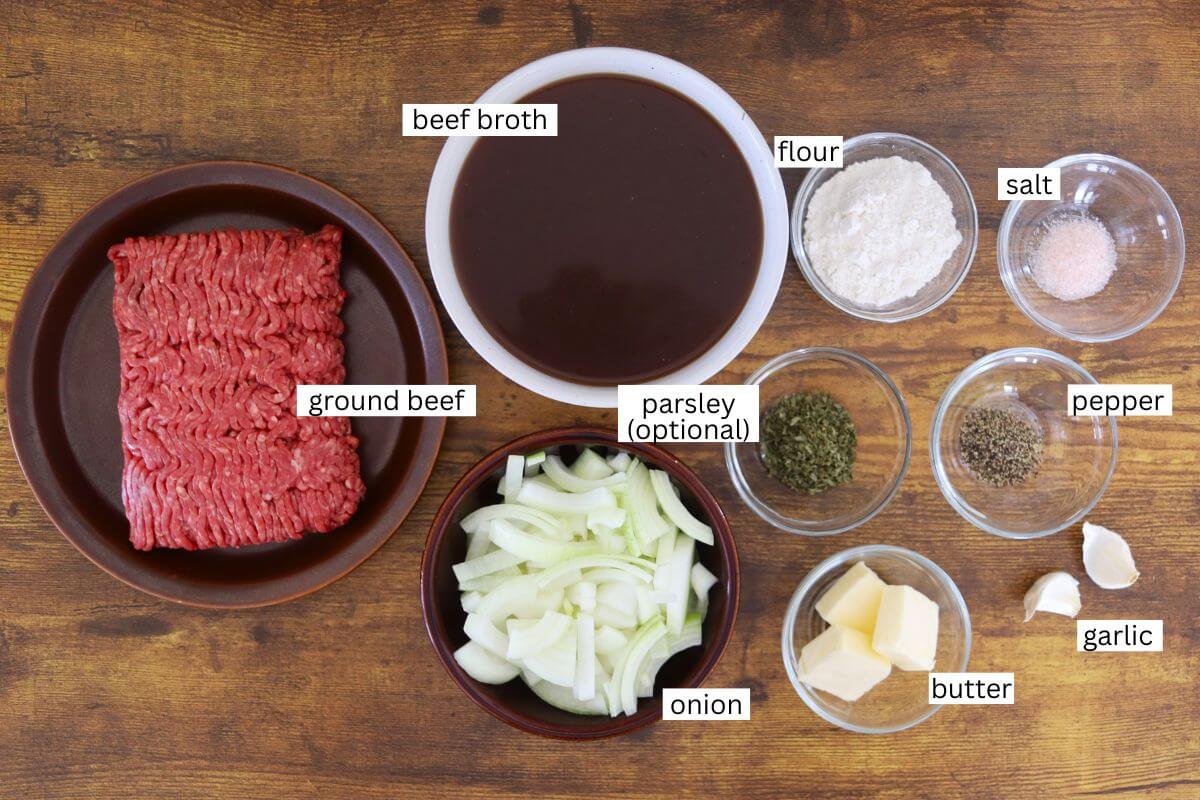 Labeled ingredients for old-fashioned ground beef and gravy, including ground beef, beef broth, onion, butter, flour, garlic, salt, pepper, and optional parsley arranged on a wooden surface.