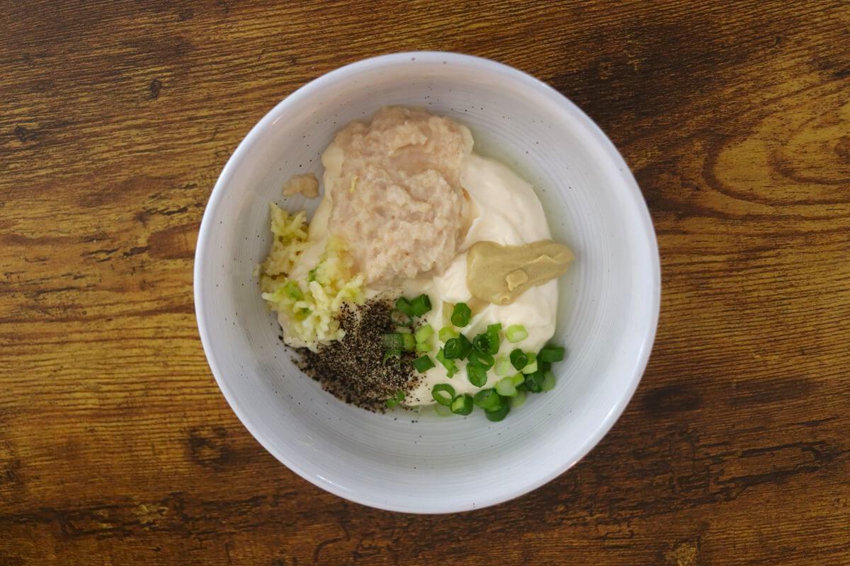 A white bowl with mayonnaise, lemon juice, horseradish, pressed garlic, dijon mustard, pepper, and scallions in it.