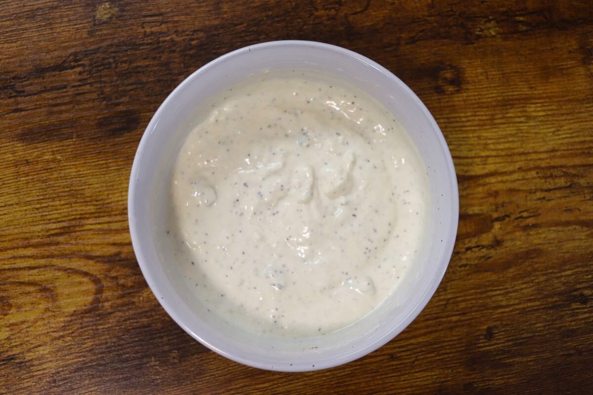 White sauce in a white bowl, with seasoning visible in it, sitting on a wooden table.