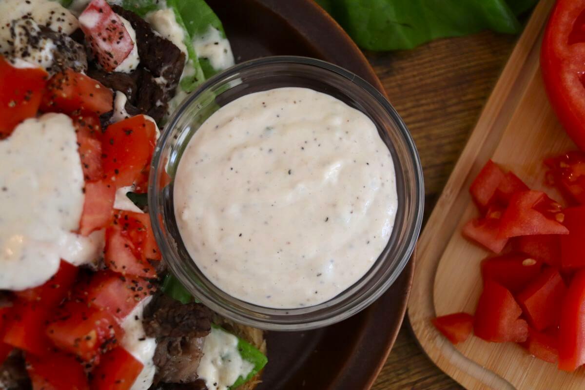 A small glass bowl filled with creamy white dressing sits on a plate next to chopped tomatoes, lettuce, and pieces of beef, with extra diced tomatoes on a wooden board nearby.