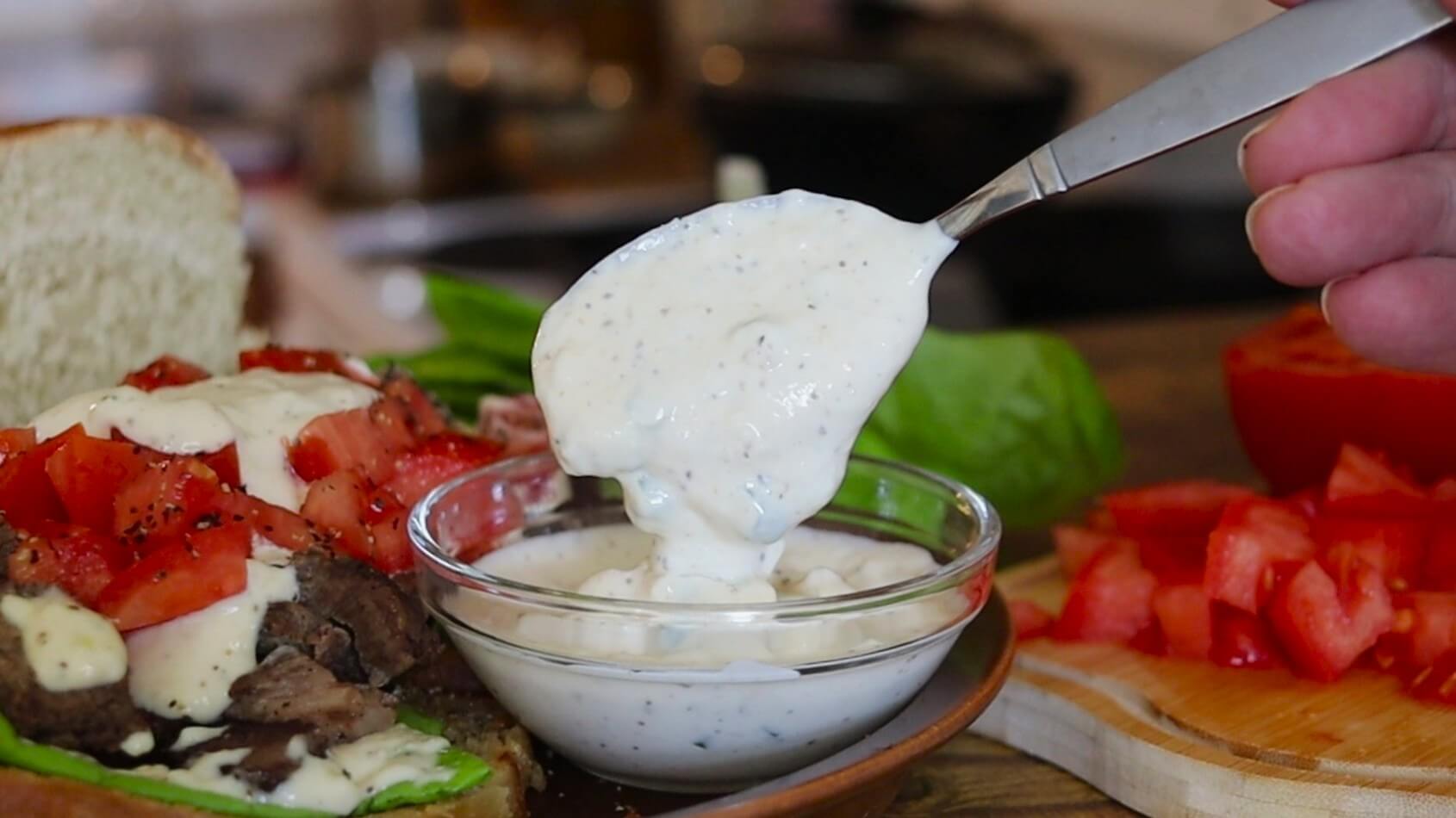 A spoon is held over a bowl of creamy white sauce, with the sauce dripping into the bowl, next to an open-faced sandwich with chopped tomatoes and lettuce. Sliced tomatoes and lettuce are visible in the background.