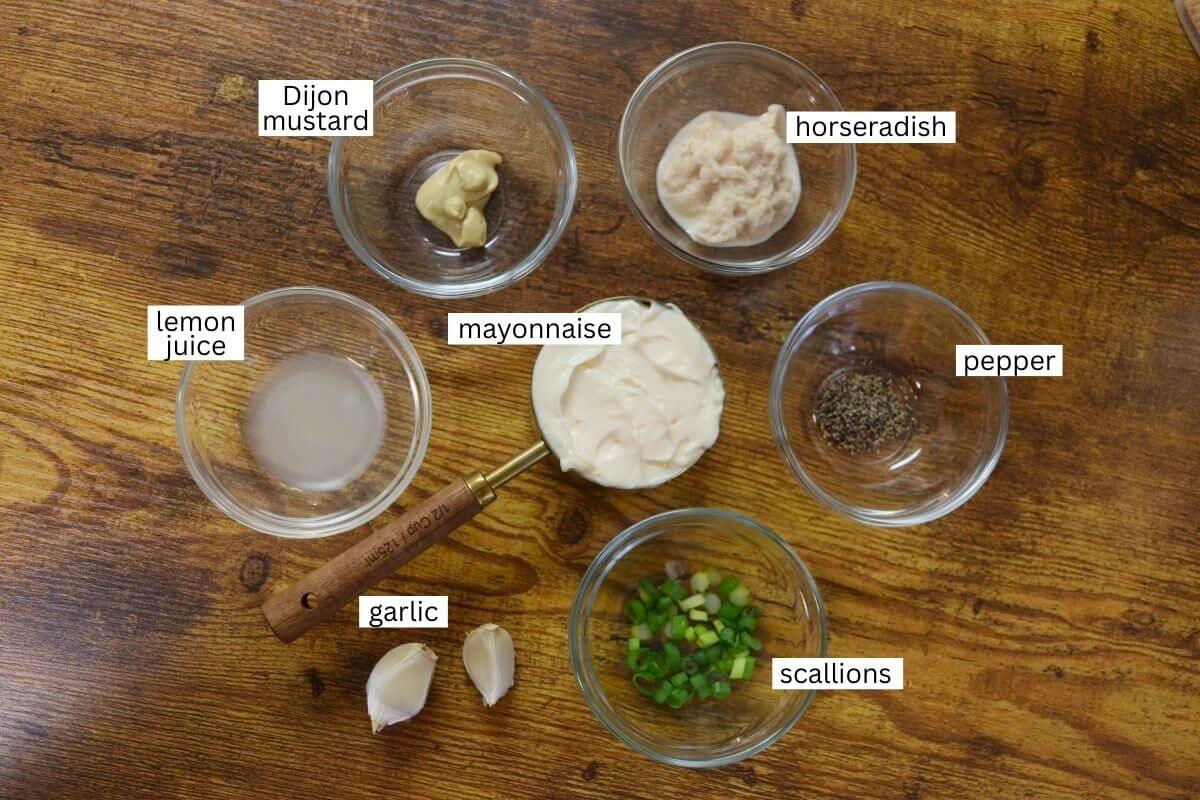 Six small bowls arranged in a circle on a wooden surface, each containing ingredients labeled as Dijon mustard, horseradish, mayonnaise, black pepper, lemon juice, garlic cloves, and chopped scallions.