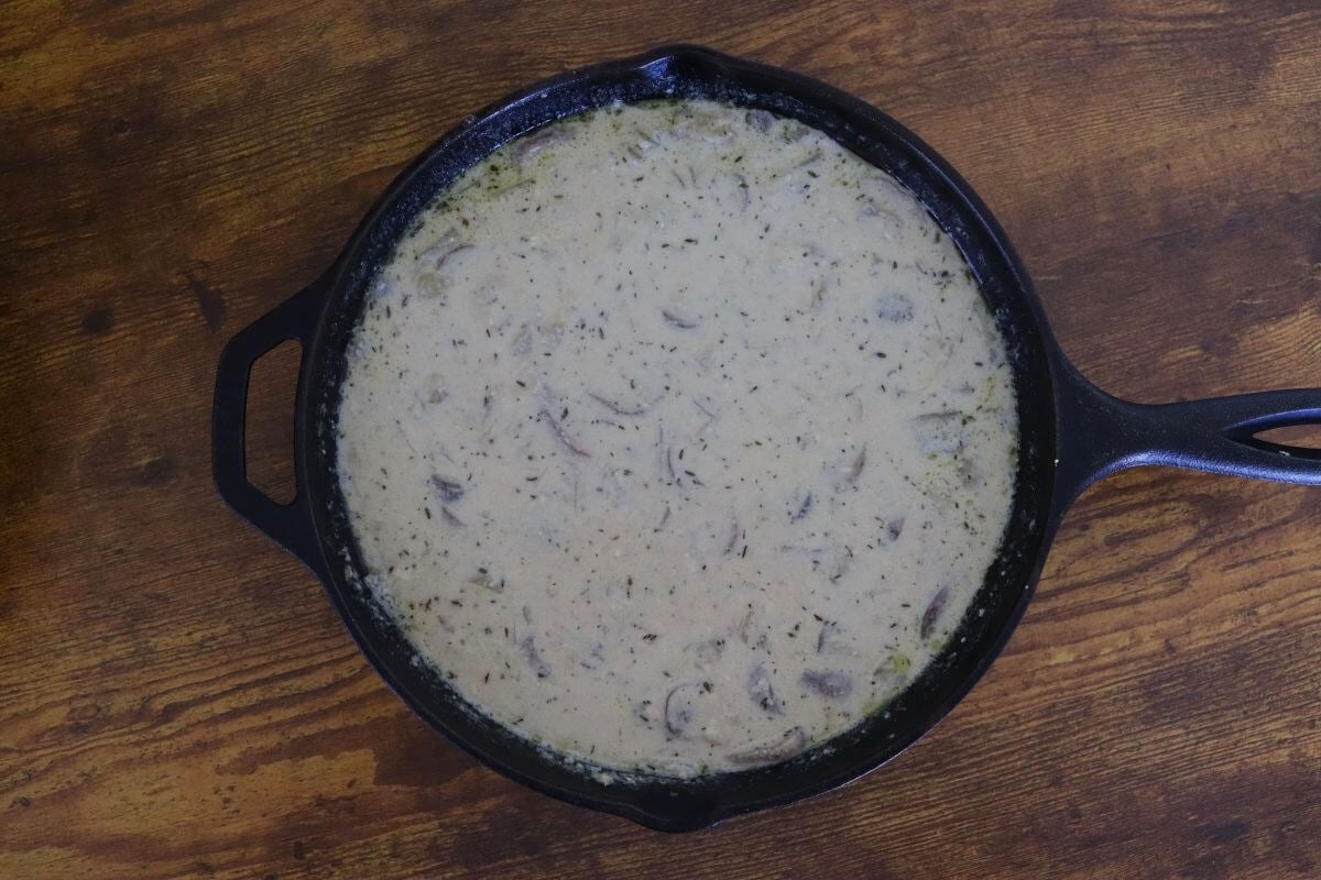 Creamy mushroom sauce with visible herbs resting in a cast iron skillet on a wooden surface.