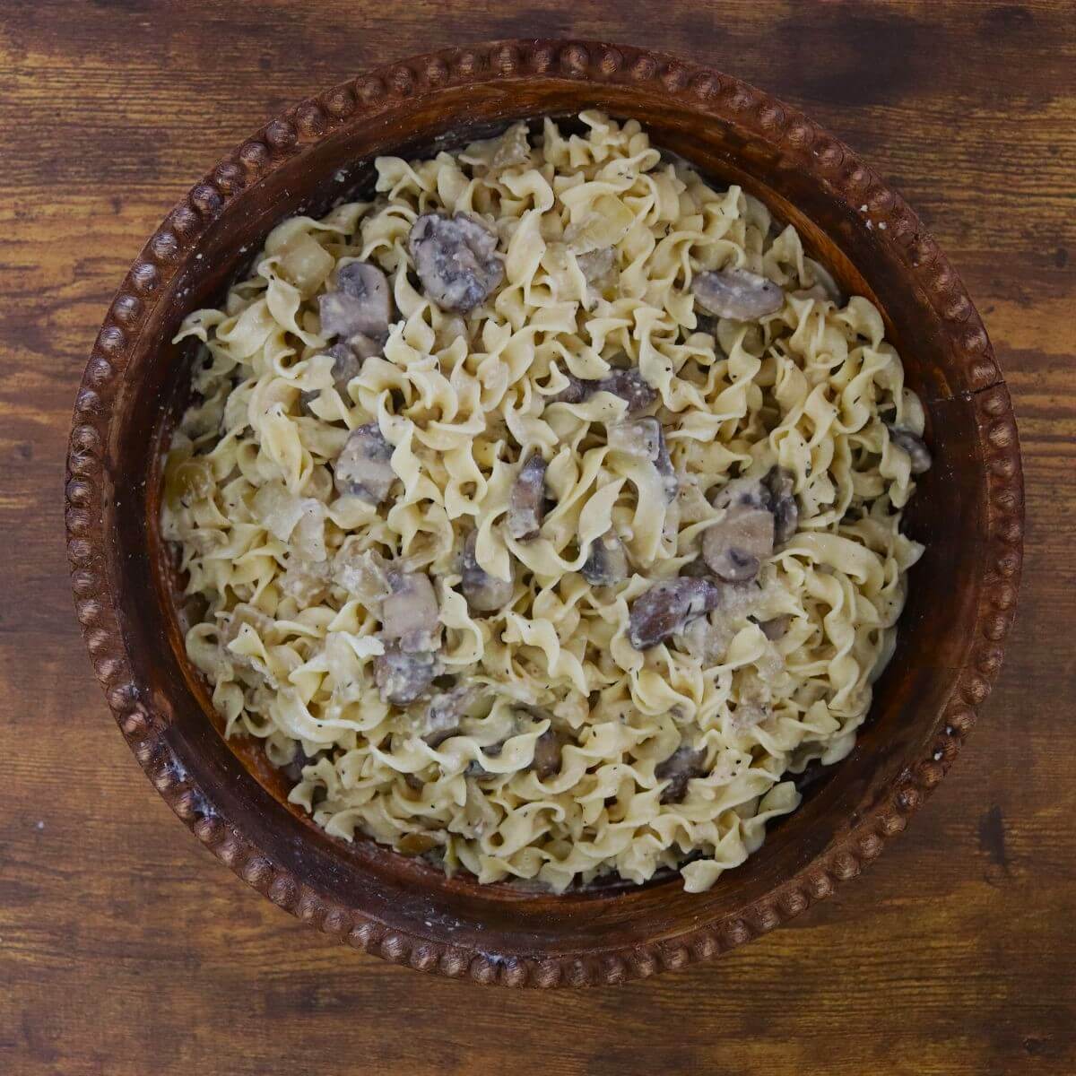 Egg noodles coated in a creamy mushroom sauce with visible slices of sautéed mushrooms, served in a rustic wooden bowl on a table.