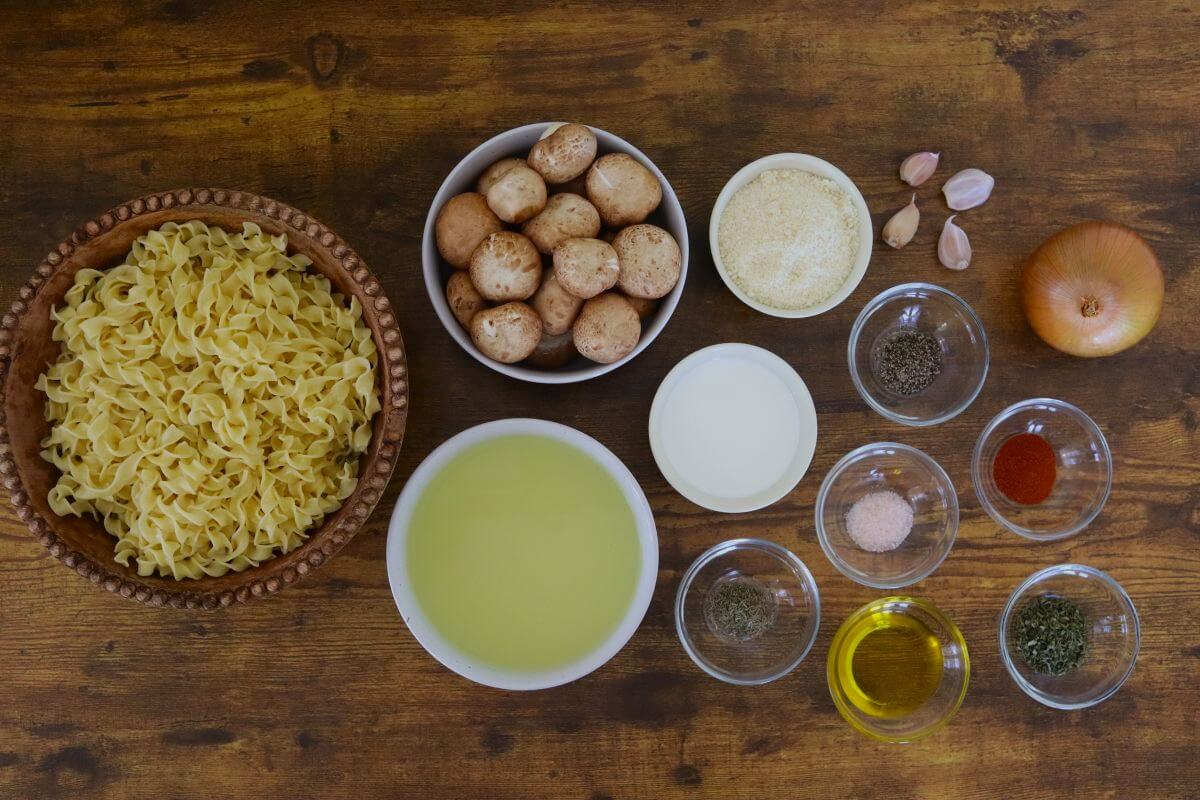 Uncooked egg noodles, whole mushrooms, bowls of liquid ingredients, grated cheese, spices, garlic cloves, and a whole onion arranged on a wooden table.