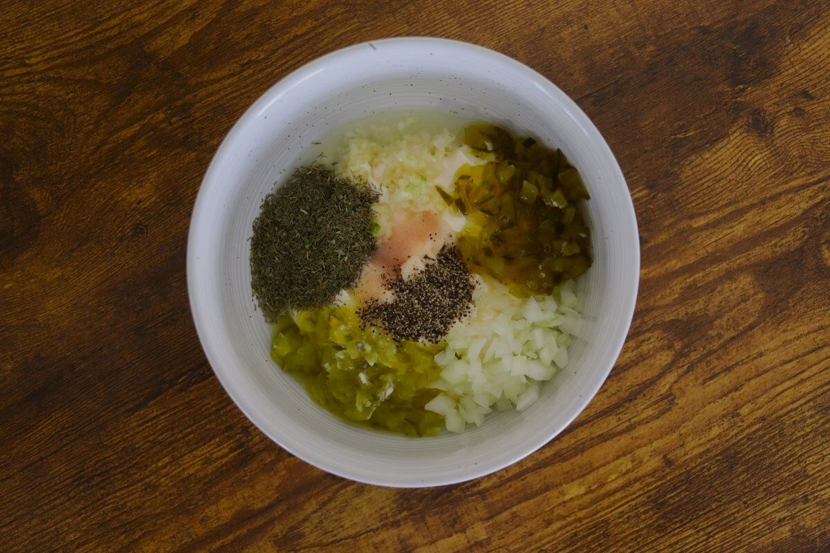 Mayonnaise, lemon juice, pressed garlic, sweet relish, dill relish, salt, pepper, and chopped onion in a white bowl.