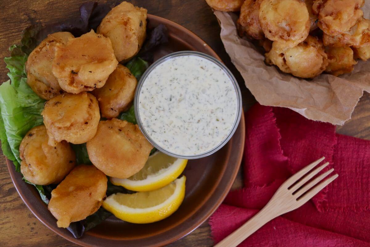 A plate of golden fried battered cod bites with lemon wedges, leafy greens, and a cup of old fashioned southern tartar sauce. A pile of more cod bites sits on brown paper, next to a wooden fork and red napkin.