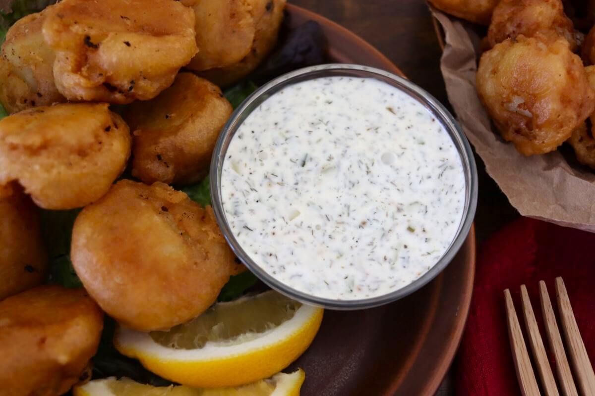 A bowl of creamy tartar sauce sits on a plate next to golden-brown fried fish pieces and lemon wedges, with more fried food and a red napkin visible in the background.