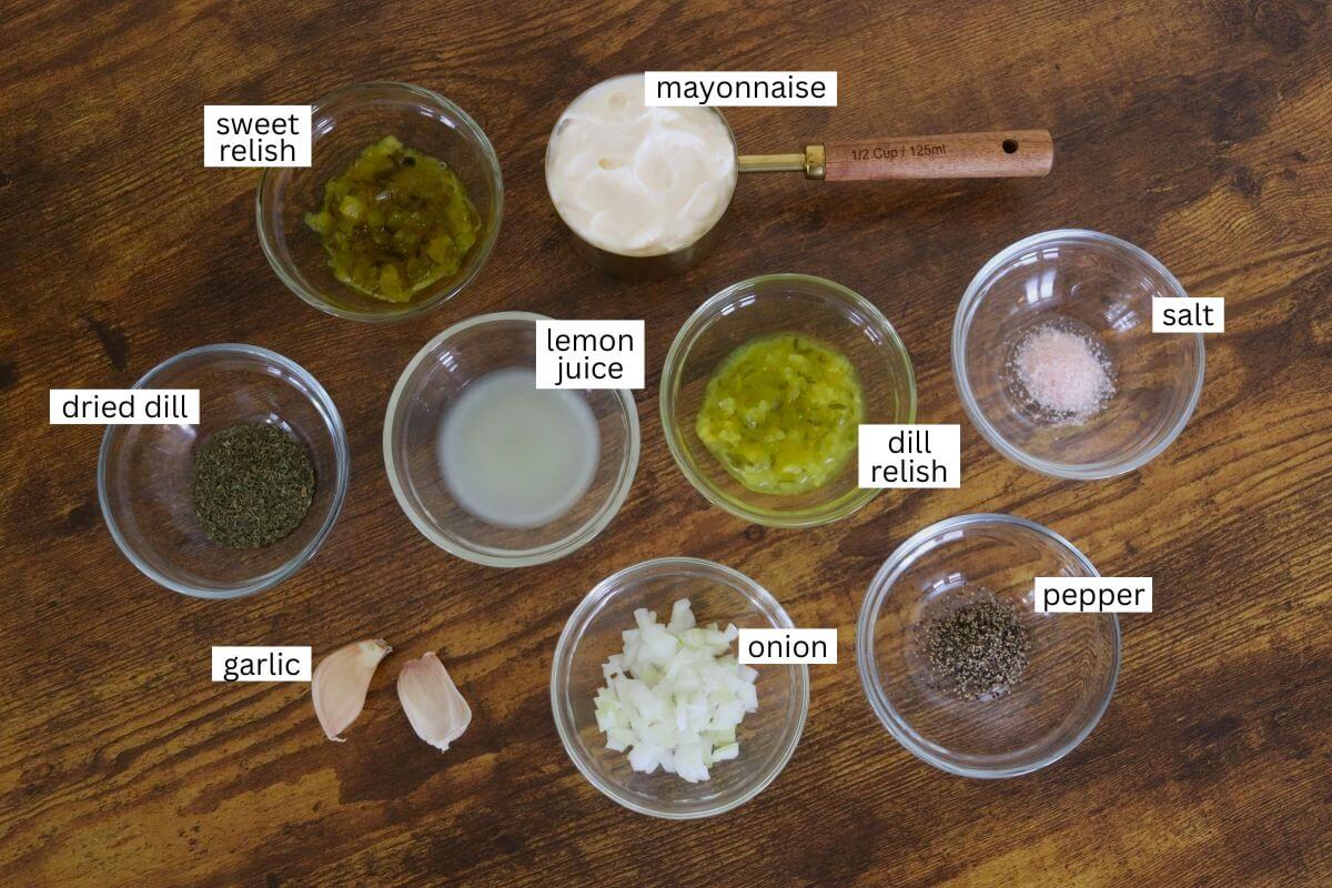 Nine labeled bowls and ingredients on a wooden surface: sweet relish, mayonnaise, dried dill, lemon juice, dill relish, salt, garlic cloves, chopped onion, and pepper.