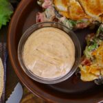 Light orange sauce in a small dish on a plate with a grilled sandwich visible to the side of it.
