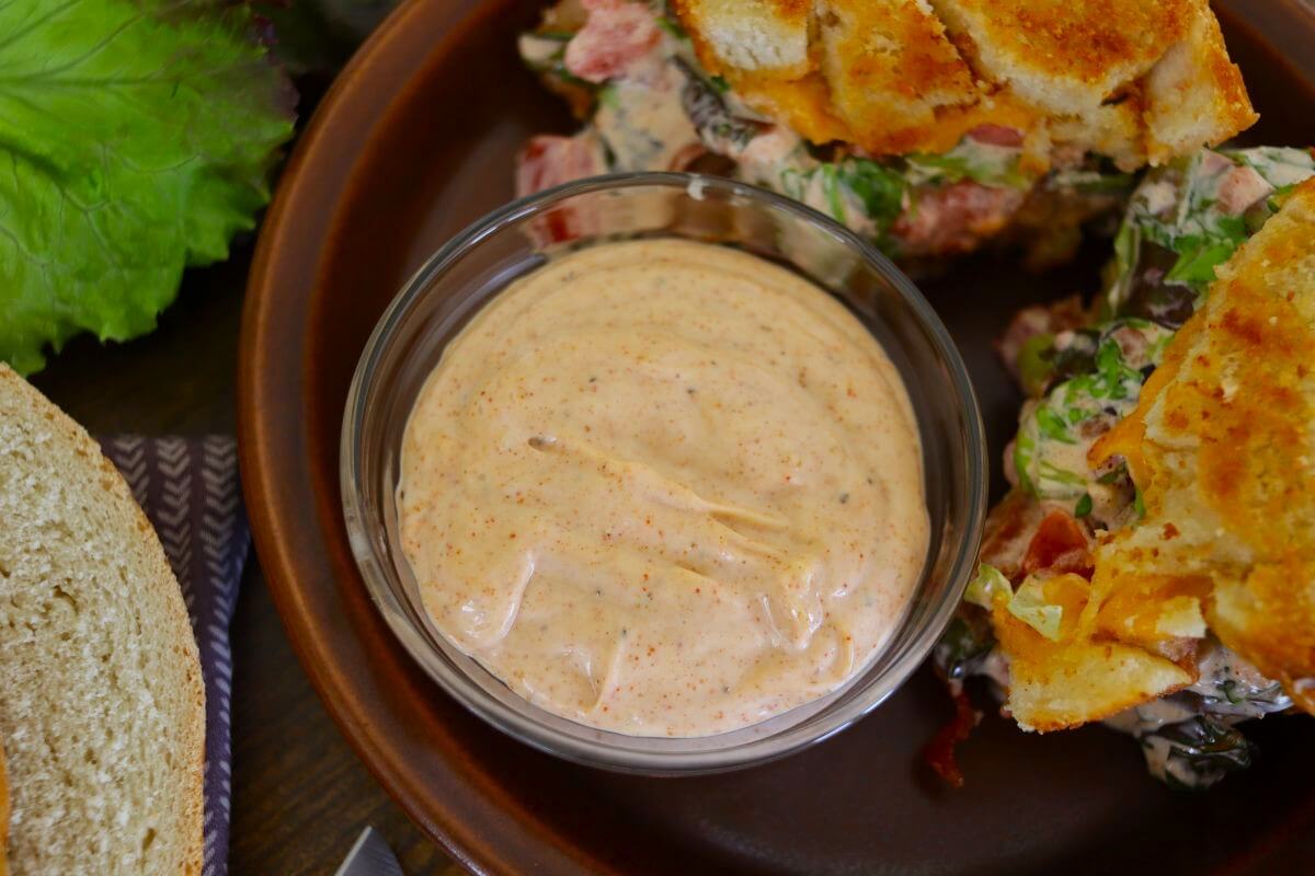 A small glass bowl filled with creamy, light orange sauce sits on a brown plate next to a sandwich with lettuce and tomato. A slice of bread and leafy greens are visible nearby.