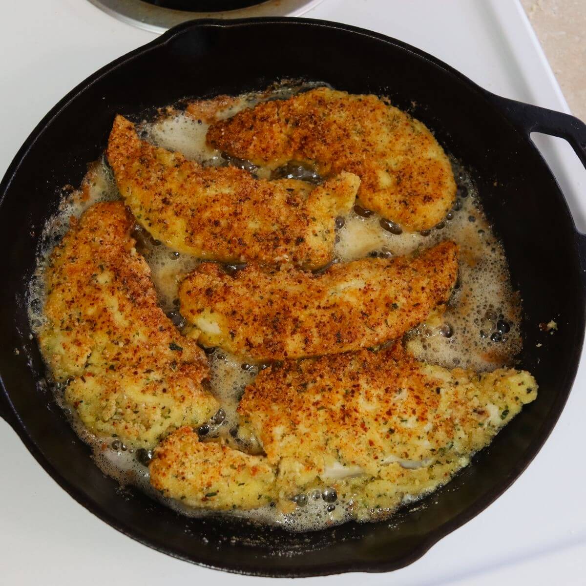 Almond crusted chicken tenders cooking in bubbling oil inside a cast iron skillet.