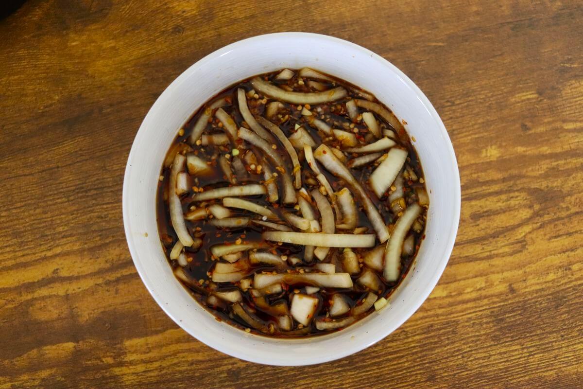 White bowl with soy-based marinade, sliced onions, and red pepper flakes on wooden surface.