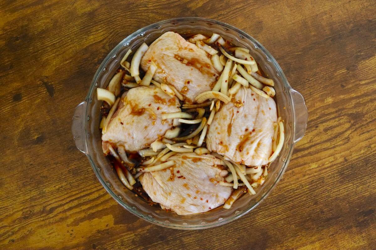 Glass dish with raw chicken thighs marinating in soy sauce mixture with sliced onions.