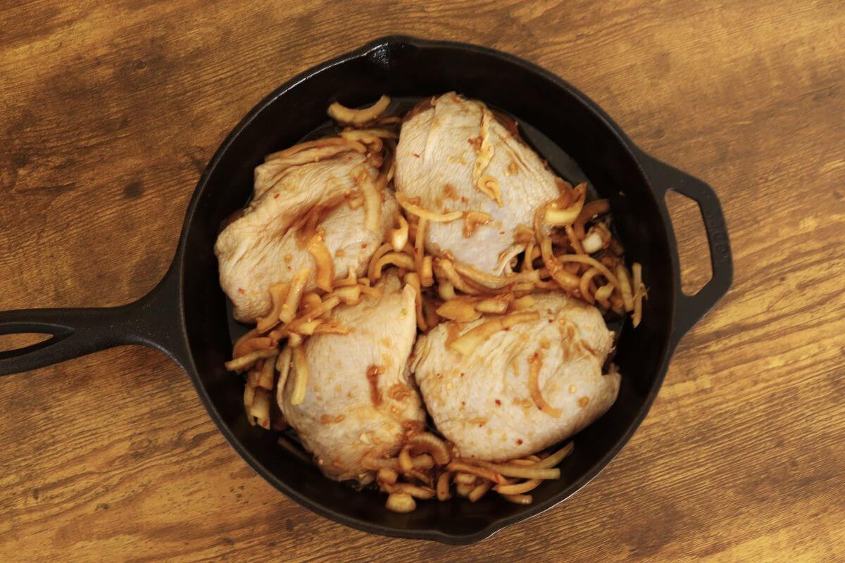 Cast iron skillet with raw marinated chicken thighs and sliced onions.