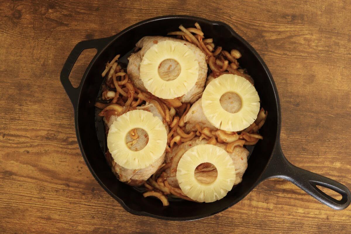 Cast iron skillet with marinated chicken thighs topped with pineapple rings and sliced onions.