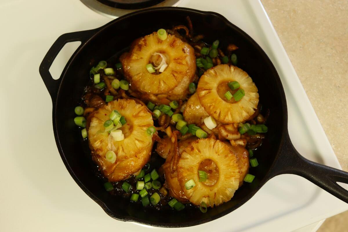 Skillet of glazed chicken thighs with pineapple slices and green onions on white stovetop.