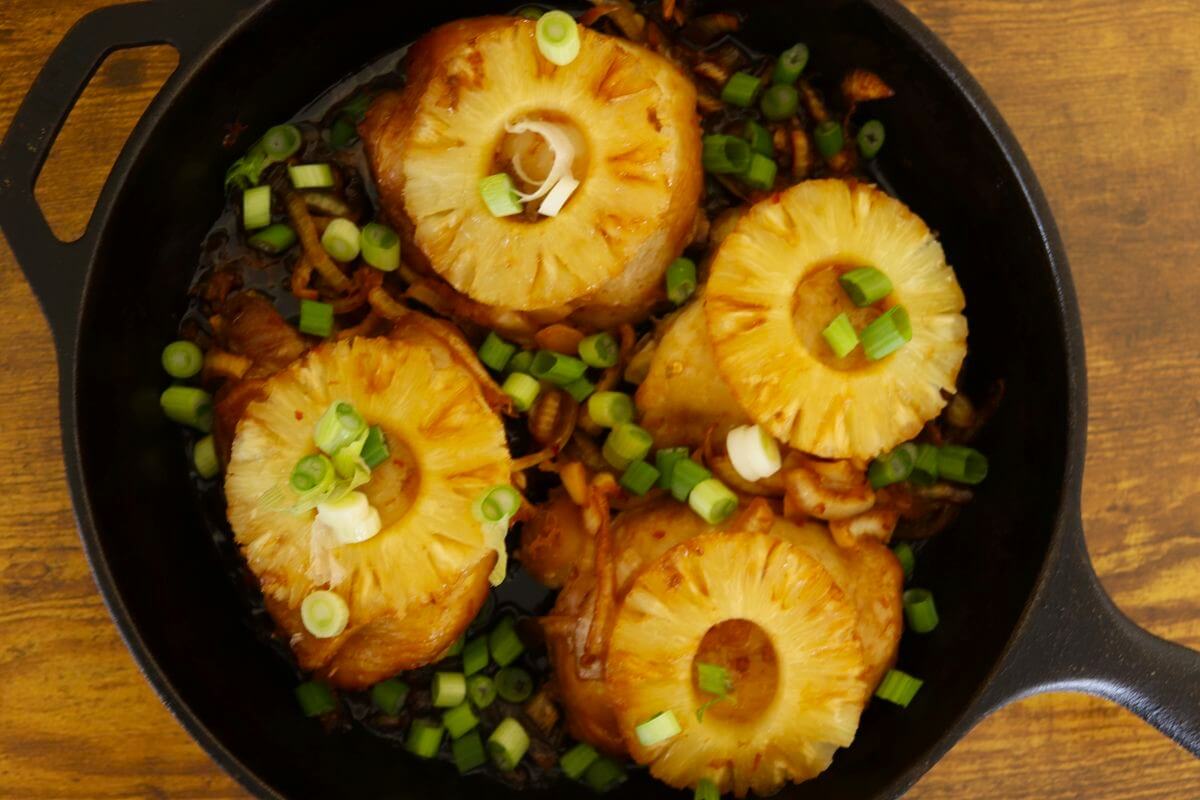 Cast iron skillet with baked Hawaiian BBQ chicken thighs topped with pineapple rings and chopped scallions.