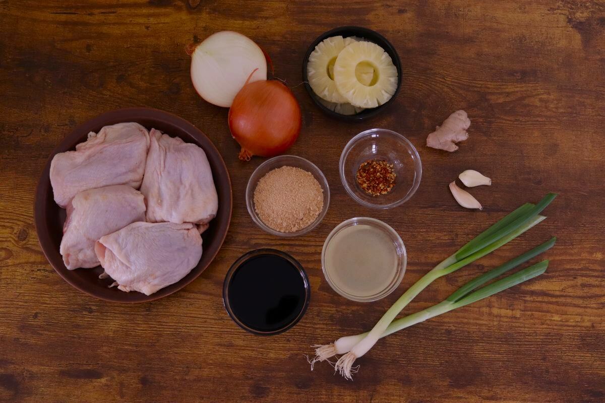 Raw chicken thighs with pineapple rings, onion, ginger, garlic, soy sauce, and brown sugar on wooden surface.