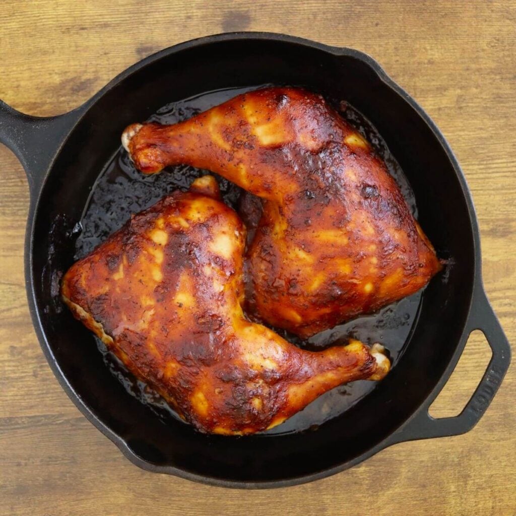 BBQ chicken leg quarters with browned, sticky barbecue sauce in a cast iron skillet.