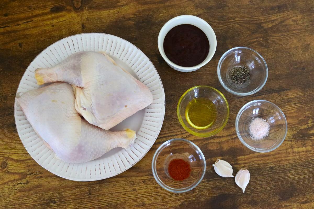 Raw chicken leg quarters with BBQ sauce, olive oil, garlic, salt, pepper, and smoked paprika arranged on a wooden surface.