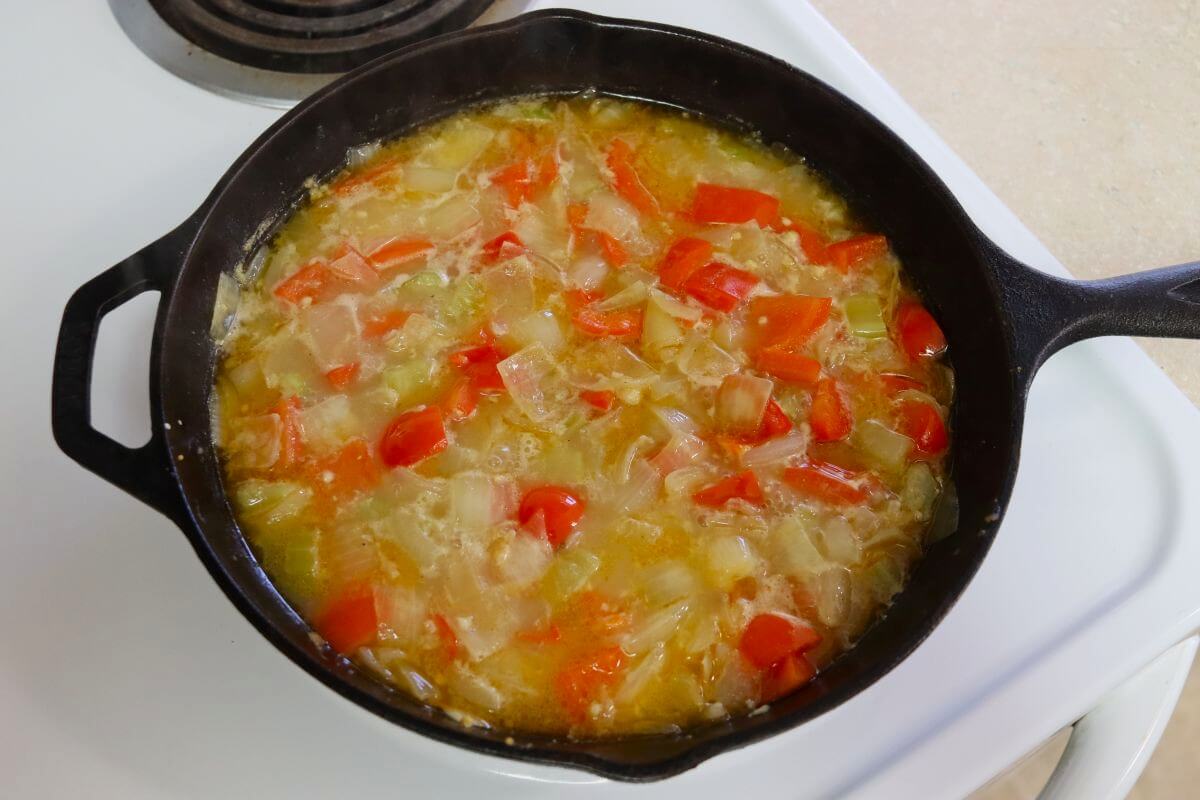 Chicken broth simmering with chopped onion, bell pepper, and celery in a cast iron skillet.