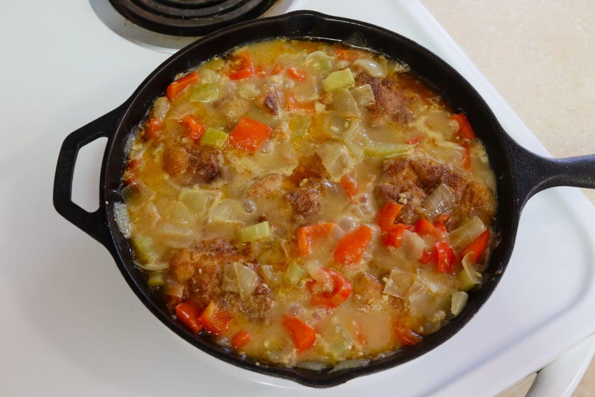 Chicken thighs simmering in chicken broth with onions, bell peppers, and celery in a cast iron skillet.