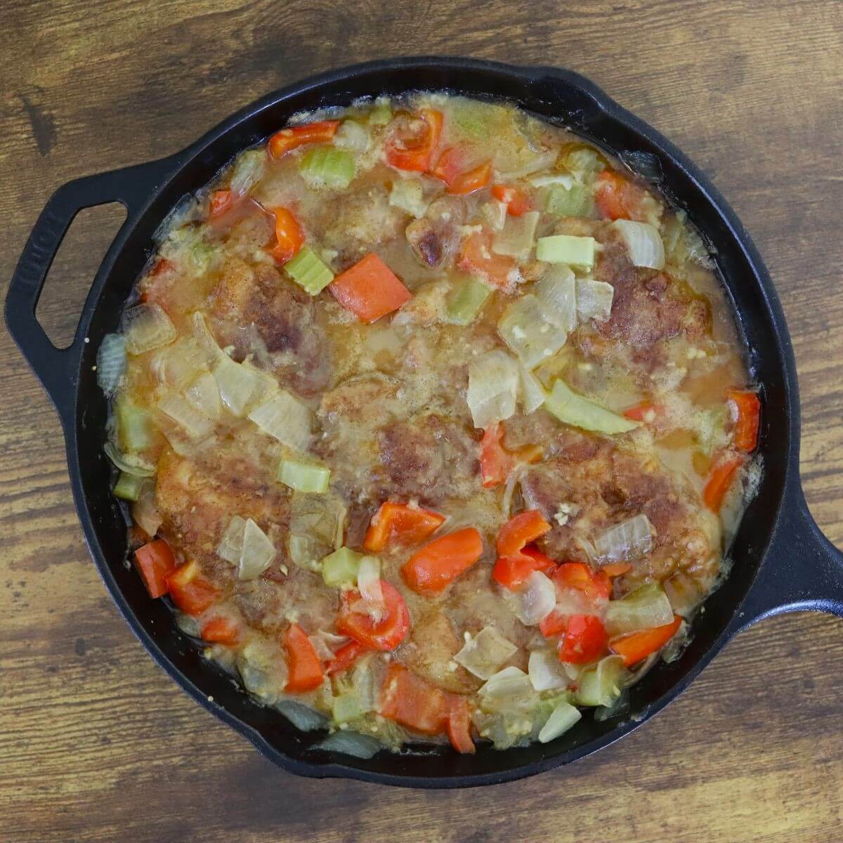 Chicken thighs in a cast iron skillet with onions, bell peppers, and celery in a light broth.