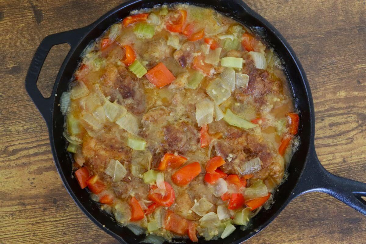 Cast iron skillet filled with chicken thighs simmering in broth with onions, bell peppers, and celery.